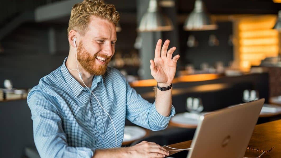 Young man sitting at his laptop in his kitchen. He's laughing and waving at someone in a video call.