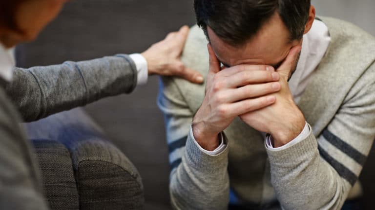 Sad or distressed middle aged man sitting, holding his head in his hands, being comforted by another man who put his hand on this man's shoulder